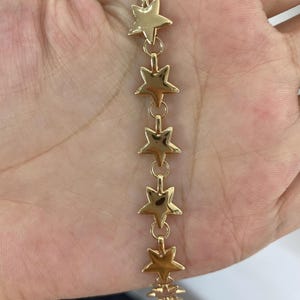 Puede incluir: Una pulsera de cadena en tono dorado que presenta una serie de dijes de estrellas de cinco puntas pulidas, unidos en una fila vertical. Cada estrella está conectada por un pequeño anillo circular, creando un diseño continuo y uniforme.