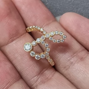 Op de afbeelding: Een gouden ring met een doorlopend, open ontwerp bestaande uit meerdere kleine, ronde, heldere stenen in een zetting. De stenen zijn gerangschikt in een lusvormig cijfer-acht patroon dat om de vinger wikkelt, met één grotere steen.