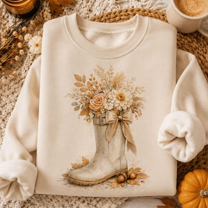 May include: A cream-coloured crewneck sweatshirt featuring a graphic print of a light-coloured rain boot filled with a bouquet of dried flowers, leaves, and grasses, accented with a large tied ribbon bow and scattered autumn leaves at the base.