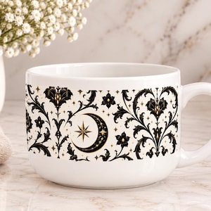 Op de afbeelding: Een witte keramische mok met een zwart en goud decoratief patroon van krullende bloemranken, sterren en een halve maan. Het ontwerp loopt rond het midden van de mok, die een brede, ronde vorm heeft en een stevig handvat aan de rechterkant.