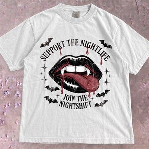 May include: A white short-sleeve t-shirt featuring a graphic of black lips with white fangs and a red tongue. The text reads SUPPORT THE NIGHTLIFE and JOIN THE NIGHTSHIFT, surrounded by small black bats and star icons with red blood drips.