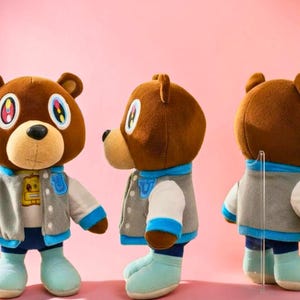 Peut inclure: Trois ours en peluche bruns portant des vestes universitaires grises et blanches avec des accents bleus, des jeans bleus et des bottes bleu clair. Les ours ont des yeux colorés et sont posés sur un fond rose.