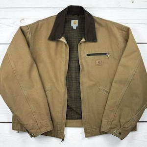 May include: A tan canvas jacket featuring a dark brown corduroy collar, a full-length front zipper, and a dark green plaid lining. The garment includes a zippered chest pocket, two lower hand pockets, and a Carhartt brand label on the chest.