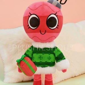 Puede incluir: Un peluche de adorno navideño con una cabeza rosa, ojos negros grandes y una boca sonriente. Lleva un suéter verde con ribete blanco y sostiene un regalo rojo. El texto "PLUSHIESHOUSE" es visible.