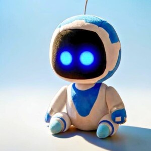 以下が含まれることがあります： 白と青のぬいぐるみロボットのおもちゃ。青く光る目と小さなアンテナが特徴です。ロボットは座っており、腕と脚が曲がっています。おもちゃはフレンドリーで漫画的なデザインです。