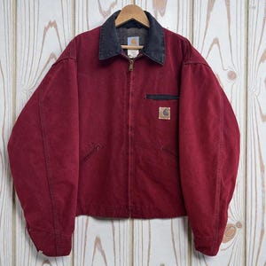 May include: A deep red canvas jacket featuring a contrasting black corduroy collar, a full-length front zipper, and two slanted lower pockets. A small rectangular Carhartt brand patch is stitched onto the left chest pocket area.