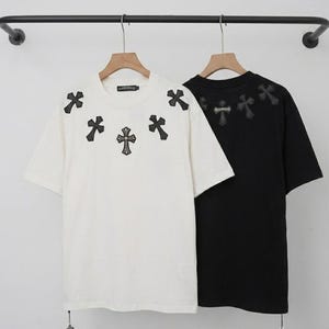 Peut inclure: Un t-shirt blanc à manches courtes et un t-shirt noir à manches courtes suspendus sur des cintres en bois. Le t-shirt blanc présente cinq écussons noirs en forme de croix autour du col, tandis que le noir affiche trois écussons similaires.