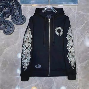 May include: A black zip-up hoodie featuring a white graphic print of ornate floral patterns along both sleeves, a circular logo on the left chest, and the letters CH near the bottom hem. The garment includes a hood with drawstrings and a silver zipper.