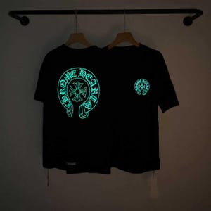 May include: A black short-sleeved t-shirt featuring a large, glowing light-blue circular graphic on the front with the text Chrome Hearts and a central cross emblem, plus a smaller matching circular logo on the upper right chest area.