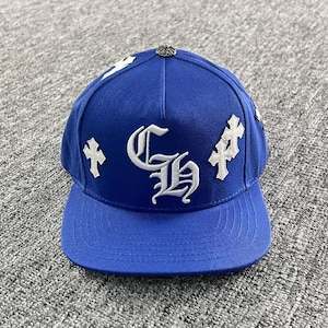 May include: A royal blue baseball cap featuring white embroidered gothic-style letters CH in the center. The cap is decorated with white cross-shaped patches on the sides and a small metallic button at the top center of the crown.
