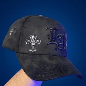 May include: A black baseball cap featuring a dark, mottled fabric texture. The side is adorned with two silver-toned metallic cross emblems. The front panel displays the letters LA in a large, dark, gothic-style font with raised embroidery.