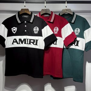 May include: Three short-sleeve polo shirts in black, burgundy, and dark green. Each shirt features a white horizontal chest stripe with the text AMIRI in black, a white collar with black stripes, three buttons, and circular AMIRI logo branding.