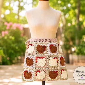 May include: A handmade crochet apron featuring a repeating pattern of chicken faces in brown, cream, and red. The apron is displayed on a mannequin and has a pink border and tie. The background is a blurred outdoor scene.