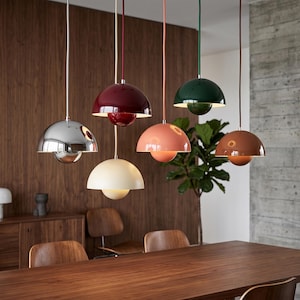 Op de afbeelding: Een verzameling hanglampen in diverse kleuren, waaronder chroom, bordeauxrood, crème, koraal, groen en bruin. Elke lamp heeft een afgeronde kap en een bolvormige onderkant, opgehangen aan het plafond met een snoer. De lampen hangen boven een houten tafel.