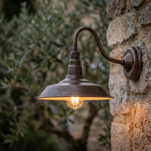 Może przedstawiać: Rustykalna, brązowa zewnętrzna lampa ścienna. Oprawa ma zakrzywione ramię i skierowany w dół klosz z widoczną żarówką w stylu Edisona. Światło jest zamontowane na kamiennej ścianie z rozmytym zielonym tłem.