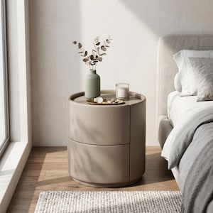 May include: A cylindrical beige bedside table with two stacked drawers. The top surface holds a light green vase containing eucalyptus stems, a small gold tray with a wristwatch, and a glass candle. The table has a smooth, matte finish.