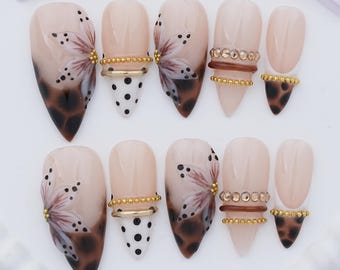 May include: A set of ten almond-shaped press-on nails featuring a nude base with varied designs including brown floral patterns, black polka dots on white, brown leopard print tips, and horizontal rows of gold beads and small brown rhinestones.