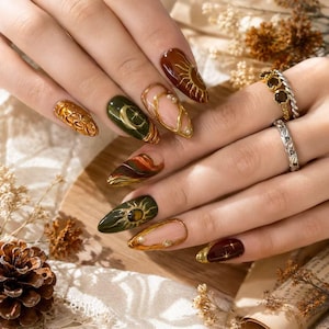May include: Almond-shaped nails featuring a mix of deep brown, forest green, and nude polish with gold metallic accents, celestial sun and moon motifs, and small embedded pearls. Two silver-toned rings are worn on the fingers, one with dark stones.