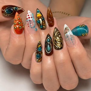 Op de afbeelding: Stiletto-vormige vingernagels met een verscheidenheid aan 3D-nailart, waaronder metalen gouden bolletjes, doorschijnende blauwe en amberkleurige cabochons, wervelende bruine texturen, witte basislagen met blauwe stippen en glanzende groenblauwe afwerkingen.