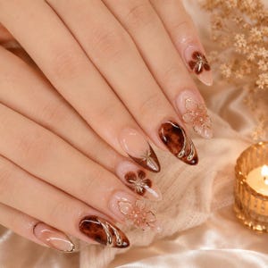 May include: Almond-shaped nails featuring a nude base with dark brown tortoiseshell patterns, gold metallic accents, and three-dimensional floral designs. The manicure includes a mix of solid brown tips, delicate gold line art, and raised flower petals.