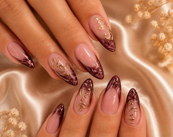 May include: Almond-shaped fingernails featuring a nude base with deep burgundy French tips. The tips have a textured, dark marble-like finish, accented with delicate, raised gold filigree swirls applied over the surface of the nails.