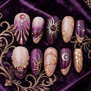 May include: A set of ten press-on nails featuring a deep purple and nude color palette. The designs include gold metallic sun and moon motifs, small white pearls, pink rhinestones, and raised gold line work on an almond-shaped nail surface.
