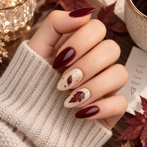 May include: Almond-shaped nails featuring a glossy deep burgundy polish on the thumb, index, and pinky fingers. The middle and ring fingers have a cream-coloured base with a dark red leaf design, speckles, and a heart icon on a nearby card.