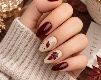 May include: Almond-shaped nails featuring a glossy deep burgundy polish on the thumb, index, and pinky fingers. The middle and ring fingers have a cream-colored base with a dark red leaf design, speckles, and a heart icon on a nearby card.