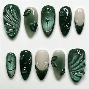 May include: A set of ten press-on nails featuring a dark forest green and cream color palette. The designs include cat-eye magnetic finishes, raised leaf-like textures, metallic star accents, and glossy spherical embellishments on almond-shaped tips.