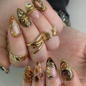 Puede incluir: Uñas en forma de almendra con intrincados detalles metálicos dorados, acentos en tono ámbar y pequeñas perlas blancas. Los dedos lucen varios anillos dorados, incluyendo un diseño prominente en forma de concha y una banda texturizada.