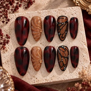 May include: A set of ten almond-shaped press-on nails featuring a dark red and black tortoiseshell pattern, shimmering gold textured accents with raised swirls, and a black butterfly wing design with small white dots on a glossy finish.