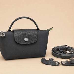 May include: A black handbag with a top handle and a fold-over flap featuring a silver-toned snap button. The bag includes a detachable shoulder strap and two small black leather side attachments with metal rings for strap connection.