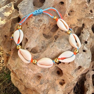 Puede incluir: Una pulsera hecha a mano con conchas blancas, cuentas doradas y cordones naranjas y azules. La pulsera se muestra sobre una superficie rocosa texturizada de color marrón claro, con luz natural que resalta los detalles.