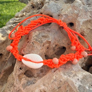 Puede incluir: Pulsera de macramé naranja brillante con una concha blanca y pequeñas cuentas de madera. La pulsera tiene un diseño tejido. Esta joya hecha a mano es ideal para la playa o el verano.