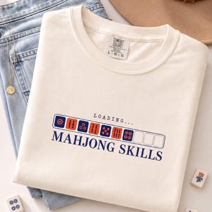 May include: A cream-colored t-shirt featuring a graphic of a loading bar composed of mahjong tiles, with the text "LOADING..." above and "MAHJONG SKILLS" below in dark blue lettering. The shirt has a Comfort Colors brand tag at the neck.