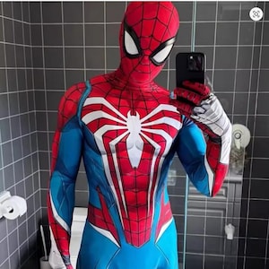 May include: A red, blue, and white Spider-Man costume with a detailed web pattern and a large white spider emblem on the chest. The costume includes a mask with white eye lenses and gloves with silver accents. The person is holding a black smartphone.