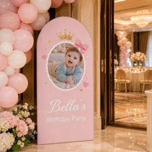 Puede incluir: Un letrero rosa alto y arqueado con una foto ovalada de un bebé en el centro. El letrero está decorado con una corona dorada, un lazo rosa y pequeños iconos de corazones. El texto "Bella's Birthday Party" aparece en letras blancas.