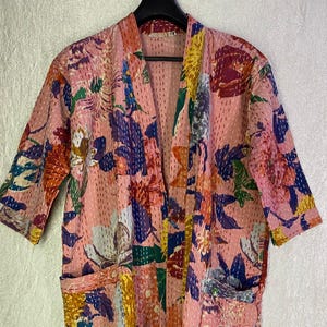 May include: A knee-length kimono-style robe in a soft pink base color featuring a vibrant, multi-colored floral print in shades of orange, blue, yellow, and white. The garment includes two front patch pockets and visible white running stitch detailing.