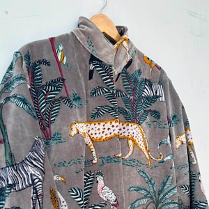 May include: A grey velvet jacket featuring a repeating jungle-themed print. The pattern includes illustrations of leopards, zebras, toucans, and cockatoos interspersed with green palm trees and tropical foliage. The jacket has a high collar and long sleeves.