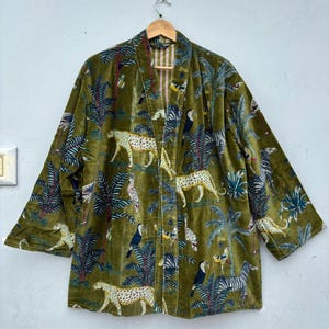 Op de afbeelding: Een olijfgroene fluwelen kimonojas met een herhalende print van luipaarden, zebra's, toekans en tropische palmbomen. Het kledingstuk heeft een open voorzijde, wijde mouwen en een zichtbare binnenvoering met verticale strepen.