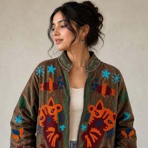May include: Brown velvet jacket with colorful embroidered cat and fish designs. The jacket features a stand-up collar and long sleeves. The embroidery includes orange, blue, and red accents. The jacket is open in the front and has pockets.