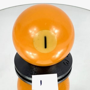 Peut inclure: Une seule boule de billard orange avec le numéro "1" jaune dessus. La boule est posée sur un support noir.