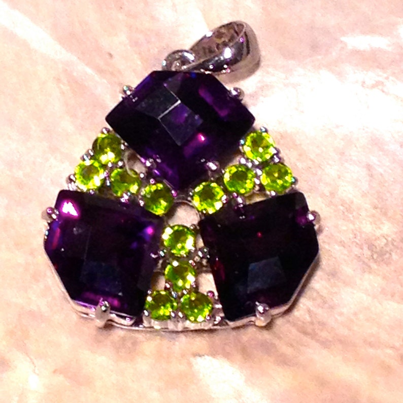 May include: A silver pendant with a geometric design featuring three square amethyst stones surrounded by smaller green peridot stones.