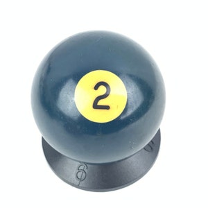 May include: A dark blue billiard ball with a yellow number "2" on it. The ball is sitting on a black base.