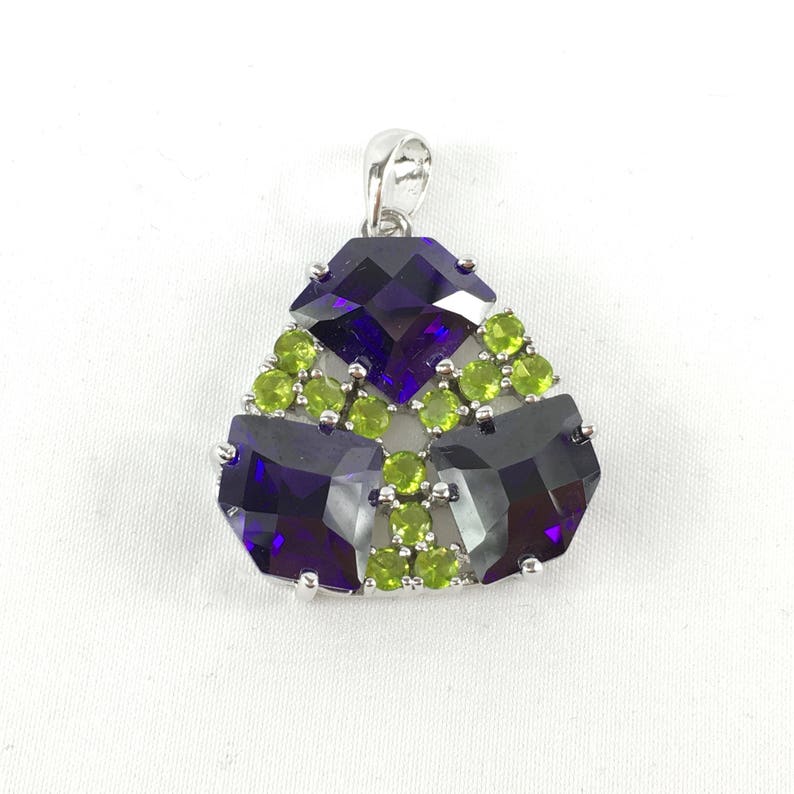 May include: A silver pendant with a geometric design featuring three large, faceted, purple amethysts surrounded by smaller, green peridot stones.