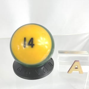 Könnte beinhalten: Eine gelbe Billardkugel mit grünem Rand und der Nummer 14 in Schwarz auf der Kugel.