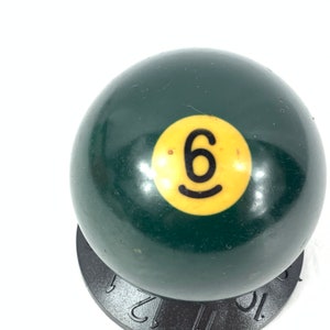 May include: A green billiard ball with a yellow number six on it. The ball is sitting on a black stand.