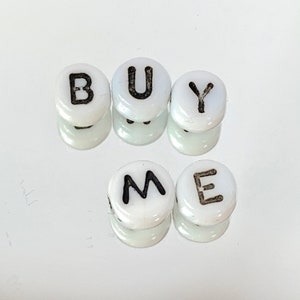May include: White round beads with black letters spelling out "BUY ME".