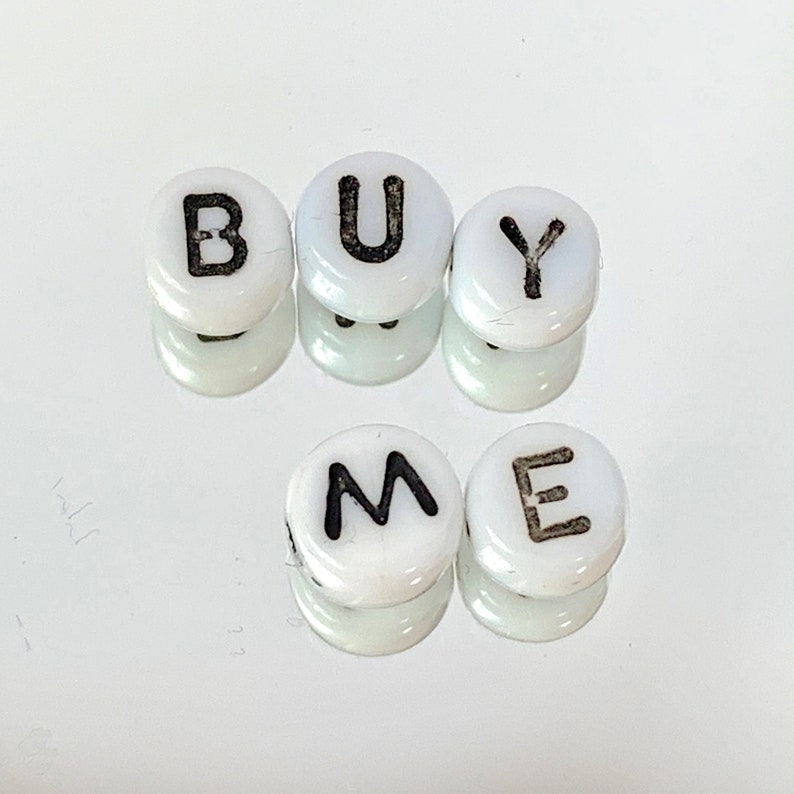 May include: White round beads with black letters spelling out "BUY ME".