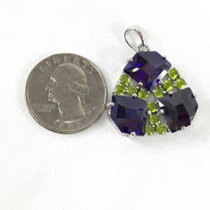 May include: A silver pendant with a triangular design featuring three large, dark purple, faceted gemstones surrounded by smaller, light green, faceted gemstones.